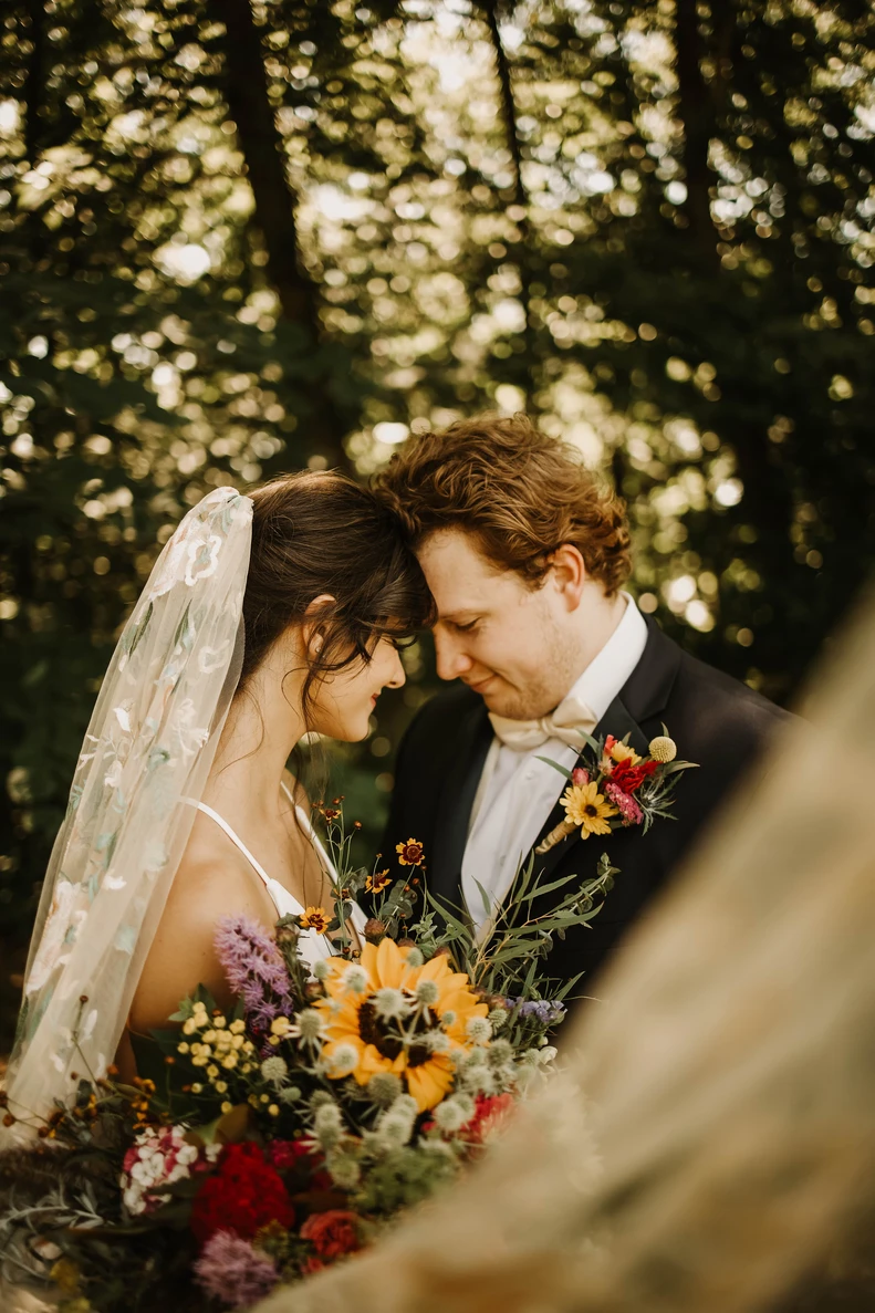 A couple leans forehead to forehead in warm forest light with a wildflower bouquet.